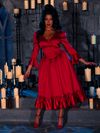 With candles casting a soft, romantic glow, a beautiful model stands in a dimly lit dungeon room, adorned in the BRAM STOKER'S DRACULA Mina Satin Bustle Dress in Blood Red.