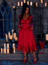 Standing amidst candles in a dimly lit dungeon room, a beautiful model showcases the BRAM STOKER'S DRACULA Mina Satin Bustle Dress in Blood Red.
