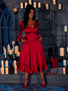 Surrounded by candles, the stunning model stands in a dimly lit dungeon room, wearing the BRAM STOKER'S DRACULA Mina Satin Bustle Dress in Blood Red.