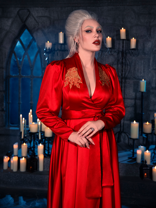 A female model strikes a pose in a candle-lit dungeon, elegantly draped in the BRAM STOKER'S DRACULA Embroidered Order of the Dragon Wrap Dress in Scarlet Red from the gothic attire label La Femme en Noir.
