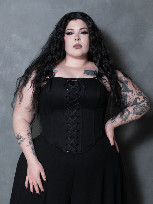 Woman wearing a black front lace up corset against a gray background. Removable straps