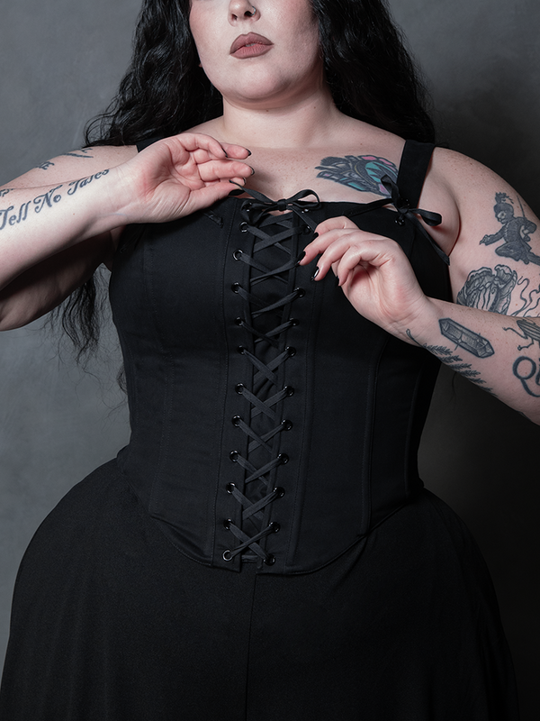 Woman wearing a black front lace up corset against a gray background. Removable straps