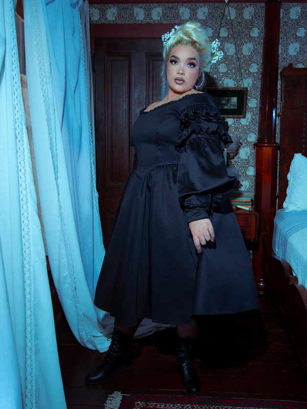A female model steps from the shadows, a striking silhouette of dark elegance in the Ellen Victorian Dress in Black.
