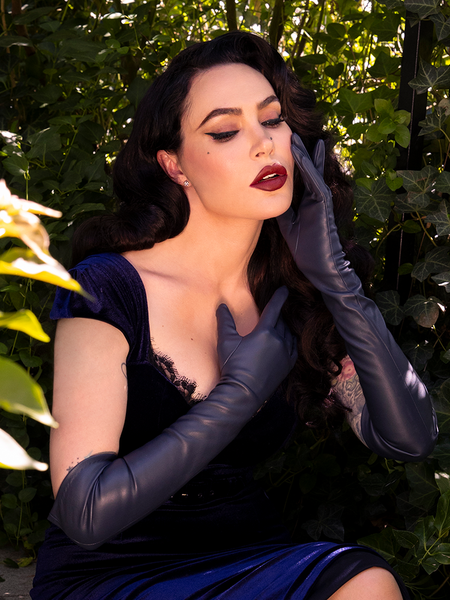 Wrapped in mystery, Micheline Pitt showcases the Faux Leather Opera Gloves in Navy from La Femme en Noir with a seductive charm in a hidden garden setting.