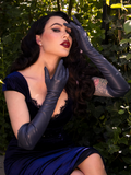 Micheline Pitt brings a touch of enchantment to a secret garden, dressed in the Navy Faux Leather Opera Gloves from the gothic brand La Femme en Noir, striking a seductive pose.
