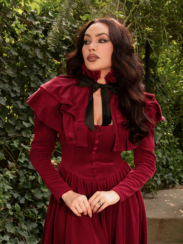 Draped in the Haunted Beauty Capelet in Crimson, Micheline Pitt stands like a gothic queen, her gaze commanding, her silhouette sculpted in silk and shadow.