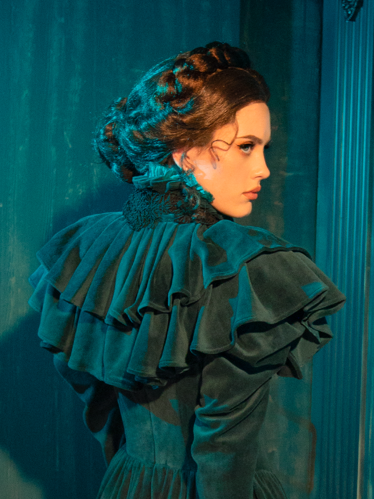 The CRIMSON PEAK™ Lady Lucille Victorian Dress with Capelet in Teal captures the allure of gothic fashion with its luxurious teal fabric, lace accents, and dramatic capelet for a truly captivating, vintage-inspired look.