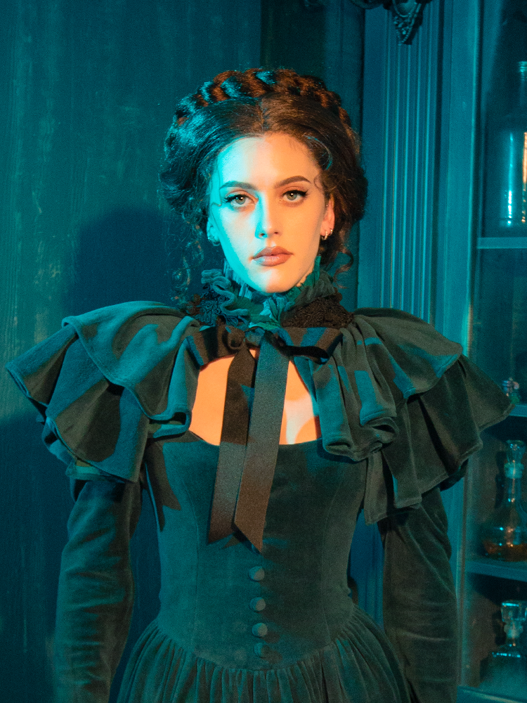 The CRIMSON PEAK™ Lady Lucille Victorian Dress in Teal is the epitome of gothic beauty, featuring a fitted bodice, flowing skirt, and a capelet that drapes elegantly from the shoulders, creating a stunning silhouette.