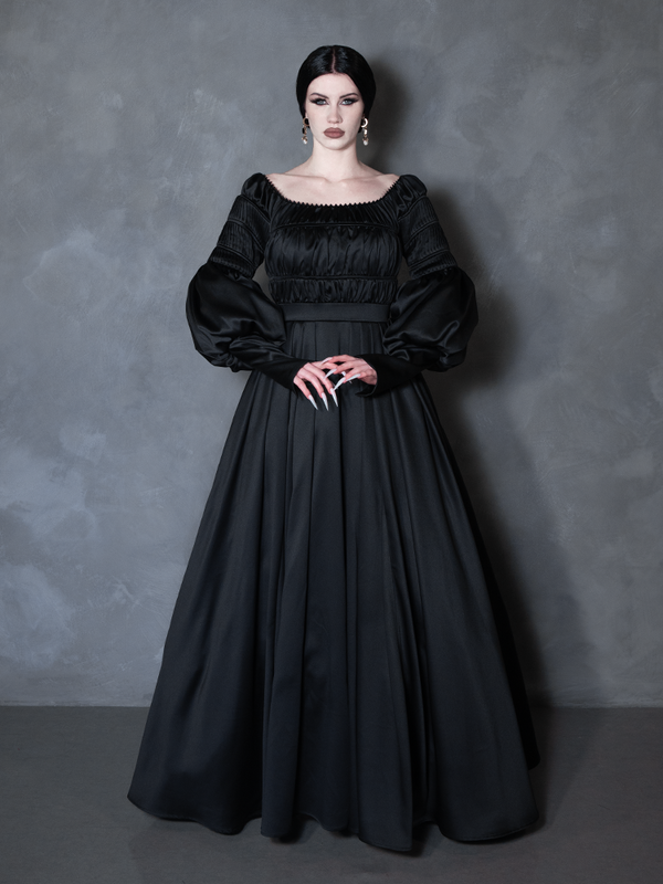 A woman modeling a long black gothic-style gown with a square neckline, smocked bodice, voluminous long sleeves, and a full skirt, posed against a textured gray studio backdrop.