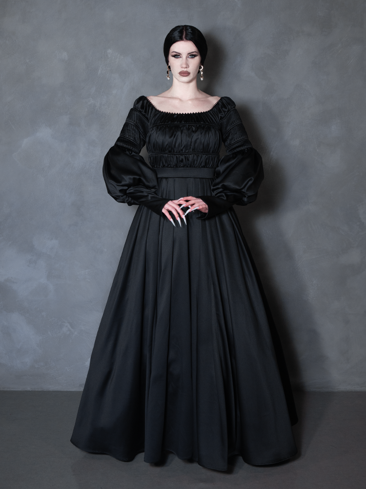 A woman modeling a long black gothic-style gown with a square neckline, smocked bodice, voluminous long sleeves, and a full skirt, posed against a textured gray studio backdrop.