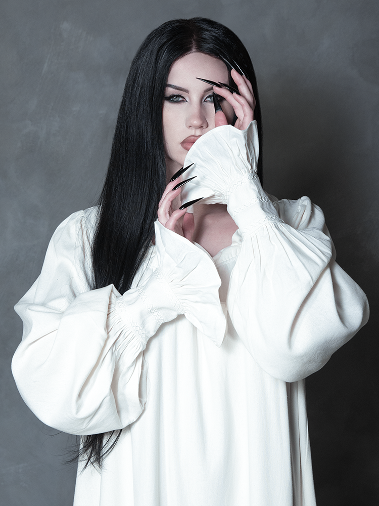 Embodying the alluring depths of dark alternative style, a female model pairs a spellbinding, shadow-kissed ivory lounge dress with an aura of pure cinematic elegance.