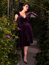 In a hidden garden setting, Micheline Pitt showcases the seductive charm of the Plum Baudelaire Swing Dress by La Femme en Noir.