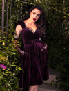 Micheline Pitt exudes allure as she seductively poses in a secluded garden, adorned in the Plum Baudelaire Swing Dress from the gothic fashion brand, La Femme en Noir.