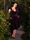 Wrapped in mystery, Micheline Pitt strikes a seductive pose in the secret garden, wearing the Baudelaire Swing Dress in Plum from La Femme en Noir.