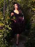 Seductively positioned amidst lush greenery, Micheline Pitt dons the Baudelaire Swing Dress in Plum from the gothic brand La Femme en Noir.