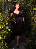In the hidden corners of a garden, Micheline Pitt captivates while wearing the Plum Baudelaire Swing Dress by La Femme en Noir.