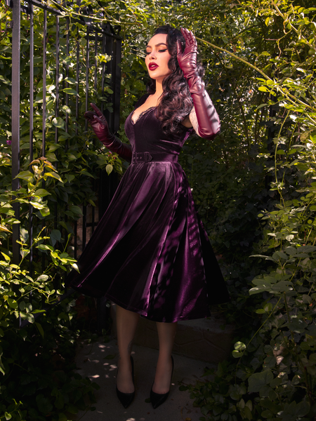 Micheline Pitt's allure is heightened in a secret garden as she wears the Baudelaire Swing Dress in Plum from the gothic clothing brand La Femme en Noir.