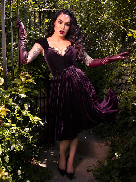 Micheline Pitt embodies gothic elegance in a hidden garden, showcasing the seductive charm of the Baudelaire Swing Dress in Plum from La Femme en Noir.
