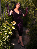 In a concealed garden setting, Micheline Pitt exudes allure while wearing the Plum Baudelaire Wiggle Dress by La Femme en Noir.