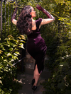 Amidst the secrecy of a garden, Micheline Pitt dons the Baudelaire Wiggle Dress in Plum from La Femme en Noir, showcasing the gothic brand's distinct style.