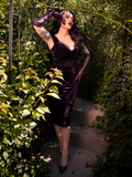 Micheline Pitt brings enchantment to a secluded garden, dressed in the Plum Baudelaire Wiggle Dress from La Femme en Noir, the gothic clothing brand.