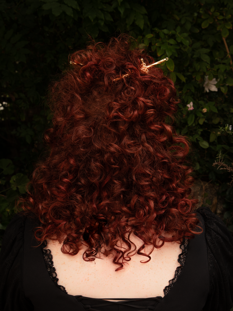 The gold sword hair pin rests atop a velvet ribbon, its metallic sheen catching candlelight. Intricate engravings along the handle whisper of forgotten battles and timeless elegance, a perfect accent for darkly romantic hairstyles.