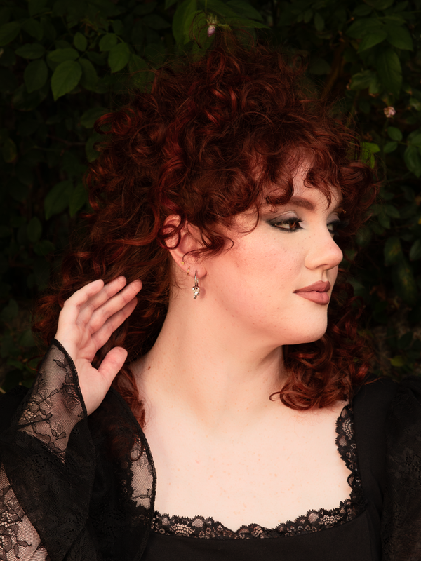 Channeling a dark, romantic allure, a female model wears silver skull hoop earrings that shimmer with an edgy anti-hero vibe, making them the ultimate statement piece for gothic style dresses.
