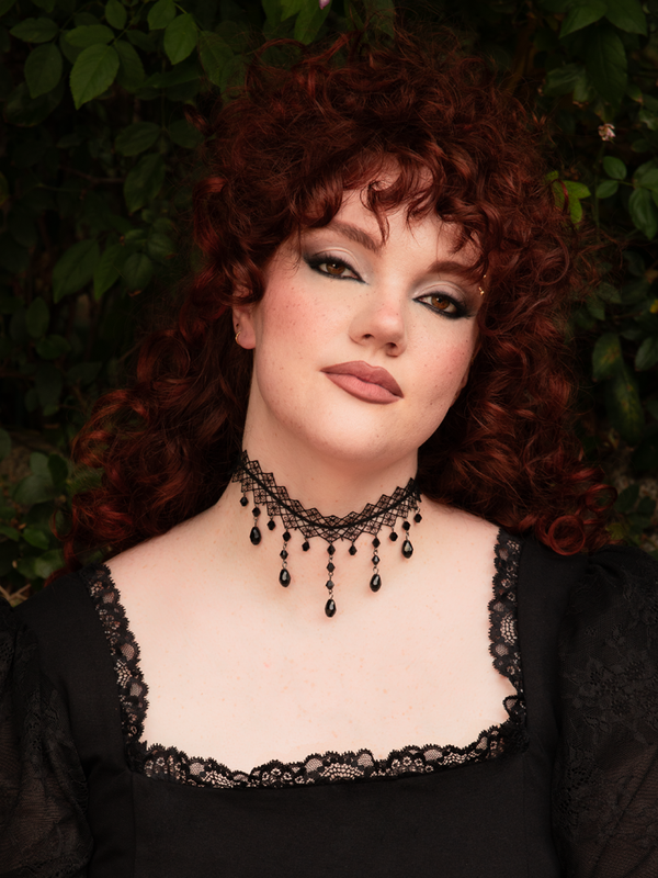 A close-up of the Woven Choker with Drop Crystals, featuring intricate black Victorian lace that clings to the neck like a dark embrace. Shimmering black glass crystals dangle from the woven trim, catching the light like frozen tears of a midnight muse.