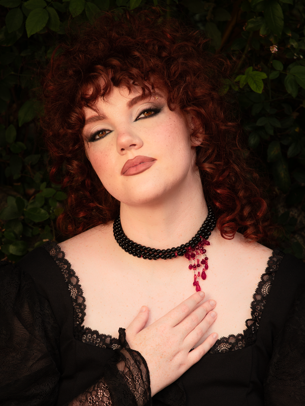 A close-up view reveals layered black strings accented with glistening red blood crystals, evoking dark romance and gothic opulence with a dramatic, vampiric edge.