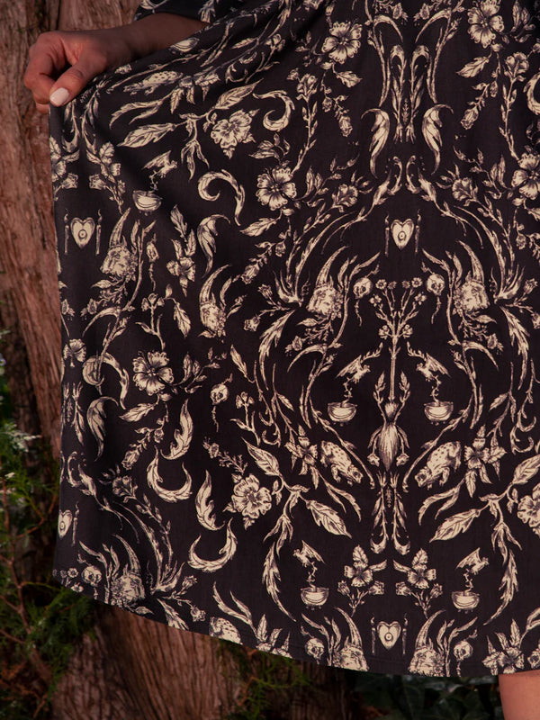 In a picturesque garden, the beautiful brunette model displays the Belladonna Maxi Dress in Cottage Witch Toile Print from the gothic clothing brand La Femme en Noir.