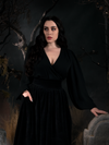 Rachel tucking one hand into her pocket and letting another rest upon a tombstone, models an all black goth glamour outfit featuring the Georgette Wrap Top in Black.