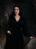Rachel tucking one hand into her pocket and letting another rest upon a tombstone, models an all black goth glamour outfit featuring the Georgette Wrap Top in Black.
