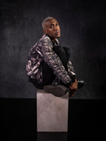 Braxton Holmes sitting on a stone colored box with his legs crossed wearing a gothic retro outfit featuring the  Alien™ Xenomorph Bomber Jacket.