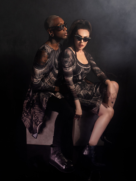 Sat on top of stone colored boxes, Micheline Pitt and Braxton Holmes pose together in items from the Alien™ collection exclusively sold by La Femme en Noir.