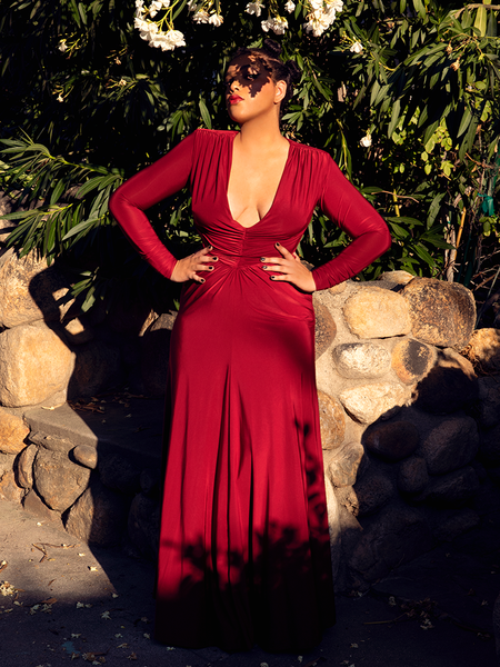 Ashleeta stands in a garden with her face shadowed by a tree while modeling the Art Deco gown in crimson.