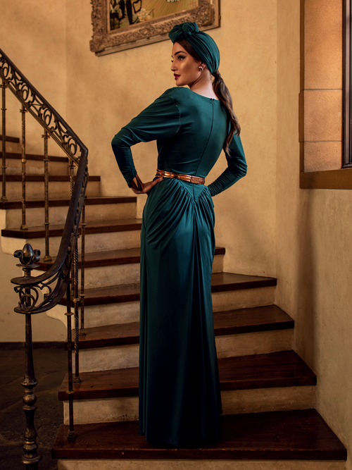 A back shot of Aliza, standing on a staircase in a gothic style home, models the Art Deco ruched gown in green from La Femme En Noir.