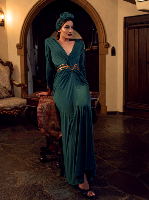 Aliza, leaning on a table in a gothic style home, models the Art Deco ruched gown in green from La Femme En Noir.