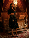 Chelsea, looking off camera while standing in a moody boudoir, models the A Spider's Kiss skirt from La Femme En Noir.