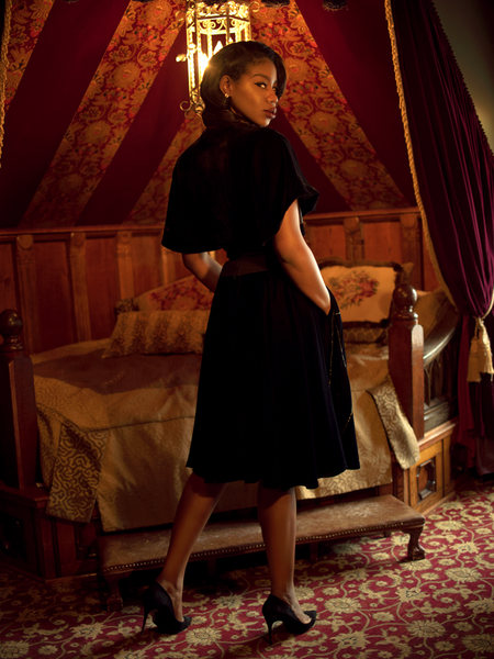 A back shot of Chelsea, standing in a moody boudoir looking over her shoulder, models the A Spider's Kiss skirt from La Femme En Noir.