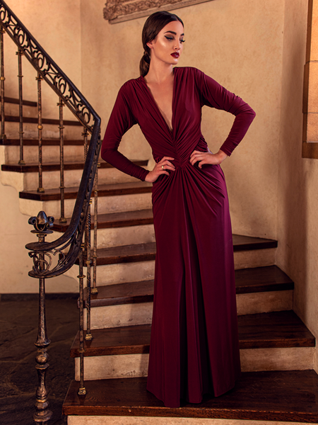 A full length shot of Aliza, standing on a staircase with her hands on her waist, models the Art Deco ruched gown in crimson from La Femme En Noir.