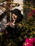 Slightly obscured by shadows, female model wears the Bolero Hat in Black to accent her gothic black dress. All items sold by goth clothing brand La Femme en Noir.