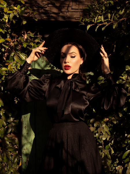 Micheline Pitt standing in front of a green garden door while wearing a gothic retro outfit including the Victorian Blouse in Black from La Femme en Noir.