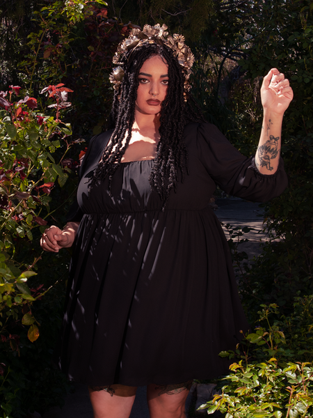 Female model poses in a garden setting while wearing the Solstice Babydoll Dress in Black Chiffon from goth dress brand and maker La Femme en Noir.