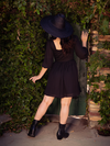 Ashley facing away from the camera to show off the back of the Solstice Babydoll Dress in Black Chiffon.