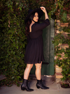Profile shot of Ashley in the Solstice Babydoll Dress in Black Chiffon standing in front of a green gate with black ankle boots on.