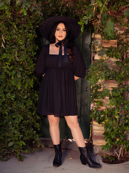 Full length shot of Ashley wearing the Solstice Babydoll Dress in Black Chiffon with matching black ankle boots.