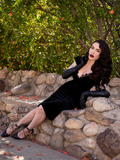 Micheline Pitt leaning on her left elbow while sitting on a stone wall wearing the Baudelaire Wiggle Dress in Black from La Femme en Noir.
