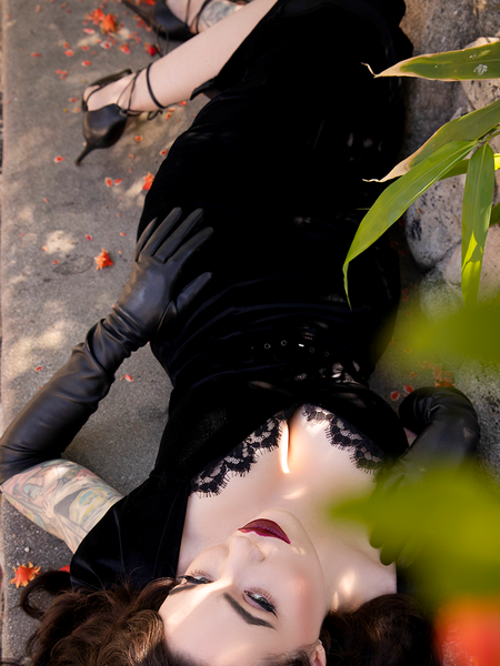 Photograph of Micheline Pitt laying down on a stone seating area in a garden setting, wears a black gothic retro style dress from La Femme en Noir.