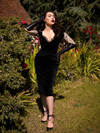 Full length shot of Micheline Pitt standing in a garden setting while wearing the Baudelaire Wiggle Dress in Black from gothic retro clothing brand La Femme en Noir.