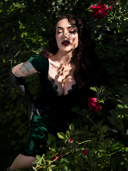 The Baudelaire Wiggle Dress in Hunter Green being worn by Micheline Pitt who poses while sitting amongst a wall of red roses. 